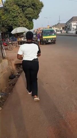 A woman with short black hair, wearing a two-tone short-sleeved shirt and black pants, walks away from the camera on a street or sidewalk. She is wearing white slip-on shoes. To her left, there is a gutter filled with trash.  To her right is a green and yellow van. Further down the street are other vehicles and pedestrians. There are also roadside vendors and parked motorcycles.