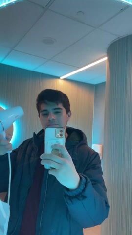 A young man is recording a selfie video in a restroom with blue-toned lighting. He is holding a white hairdryer in his left hand and an iPhone in his right hand. He begins to use the hairdryer on his hair while continuing to record the video. He pauses, looks at the camera, smiles, and the video ends. He is wearing a dark-colored puffy jacket over a maroon shirt. The background includes tiled walls and a cylindrical column with vertical ridges.