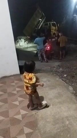 A young child wearing a yellow and orange dress stands facing away from the camera, looking towards a group of people gathered around a parked motorcycle and a yellow dump truck. The dump truck's bed is raised, appearing to be in the process of unloading. It is nighttime, and artificial lights illuminate the scene. The ground is unpaved in some areas and paved with tiles near the building on the left. The child is standing on a tiled walkway.