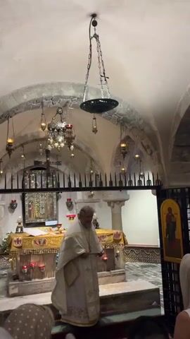 A priest in white vestments stands before an altar in a dimly lit room with stone arches and hanging lamps. He is holding his hands together at his chest and then moves them to his head. People are partially visible in the foreground.