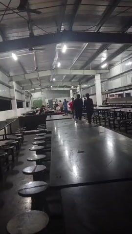 The video shows a large indoor dining hall with long tables and chairs. Several people are present, some sitting at the tables and others standing and walking around. The hall appears to be well-lit with overhead fluorescent lights. The overall atmosphere seems to be casual, possibly a cafeteria or mess hall.