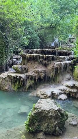 The video shows a series of cascading waterfalls flowing into a clear turquoise pool surrounded by lush green foliage and rocky cliffs. The water is transparent, revealing the rocky bottom of the pool in some areas. The overall scene is serene and natural, with the sound of running water creating a peaceful ambiance. There are no people visible in the video. The camera pans across the waterfalls and the pool, capturing the beauty of the natural landscape. The video is shot in vertical format.