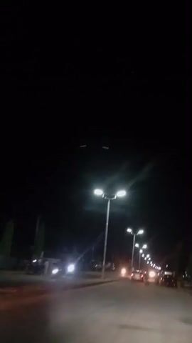 A nighttime view of a road with streetlights. The camera is moving, likely from a vehicle, as indicated by the perspective and motion blur. Multiple streetlights illuminate the scene, creating a tunnel effect down the road. Vehicles, including motorcycles and cars, are visible, some with headlights on, contributing to the illuminated path. A few figures, presumably pedestrians, can be seen near the roadside. The sky is dark, suggesting it is nighttime.