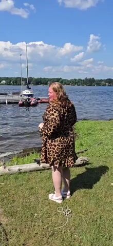 A woman in a leopard-print dress and white sandals stands on a grassy shore next to a lake. She is facing away from the camera, looking out at the water. Several boats are docked on the lake, and trees line the opposite shore. The sky is blue with scattered clouds. The woman turns her head to the left and smiles.