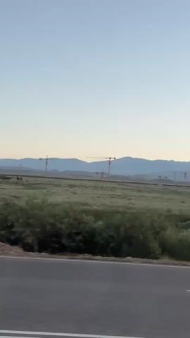 The video captures a landscape scene viewed from a moving vehicle, likely a car, driving on an asphalt road. The sky is a clear, pale blue, indicative of early morning or late evening. In the middle ground, a vast expanse of green grass and vegetation stretches out, dotted with patches of darker green shrubs and small trees. Beyond the fields, a range of mountains forms a distant, hazy backdrop. Prominently featured across the middle ground are several tall yellow construction cranes, suggesting a large-scale development project underway. These cranes are spaced out in a line, some appearing to be closer than others. The foreground shows the road and a blurred vehicle passing by, indicating the perspective is from inside another moving vehicle. The overall impression is one of a wide-open, developing landscape under a serene sky.