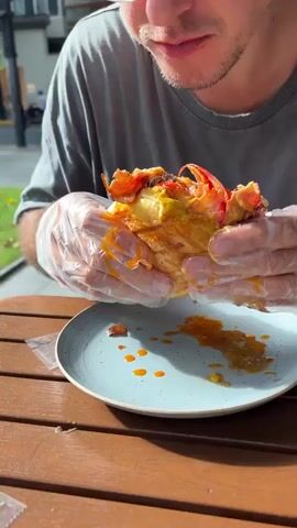 A person wearing a gray t-shirt and clear plastic gloves is eating a messy hamburger. The hamburger is large and appears to have multiple patties, cheese, and possibly bacon and tomatoes. Sauce is dripping from the hamburger onto a blue plate and the wooden table. The person is leaning over to take a bite of the hamburger. The background is blurred and appears to be outdoors with some greenery.