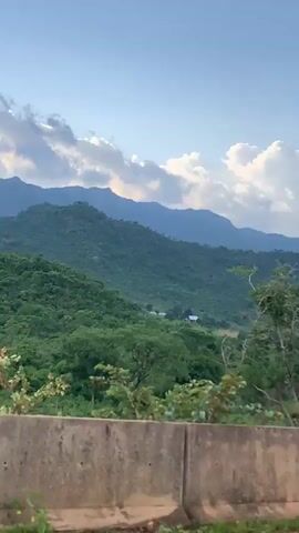 This is a vertical video clip showcasing a serene natural landscape in Nabatum, Nigeria. The scene opens with a view of lush green rolling hills and mountains in the distance, under a sky filled with scattered clouds. In the foreground, a concrete barrier runs horizontally across the bottom of the frame, with some dry brush and dirt visible below it.

As the camera pans slightly, more of the verdant landscape unfolds. The mountains appear layered, with the closest ones covered in dense foliage and the further ones receding into a hazy blue. Patches of civilization are subtly visible in the mid-ground – small white structures, likely homes or farm buildings, are nestled within the valleys and clearings amidst the greenery. The overall impression is one of rural tranquility.

The time of day appears to be late afternoon or early evening, indicated by the soft light illuminating the clouds and the gentle shadows cast across the landscape. The weather seems pleasant, with a mix of sun and clouds contributing to a dynamic sky. The abundant greenery suggests a location with sufficient rainfall, typical of a tropical or subtropical climate. The visual cues point towards a peaceful, undeveloped rural area. There is no visible text in the video. The activity is simply a slow pan of the scenic view.