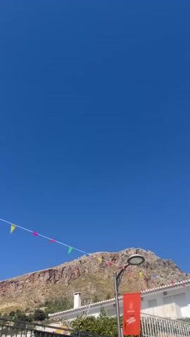 A drone is visible in the sky. The sky is clear and blue. In the lower part of the image, a mountain is visible with some buildings and a street lamp in front of it. There are also small triangular flags strung across a wire. The drone is small and appears to be moving.