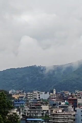 The video shows a panoramic view of a city nestled against a lush green mountain range. The sky is overcast with dense, grey clouds, and a light mist or fog seems to be clinging to the upper slopes of the mountain, obscuring some of its details. Below the mountain, a densely packed urban landscape of multi-story buildings is visible. The architecture appears to be a mix of modern and older structures, with varying colors and designs. The buildings are tightly clustered, suggesting a busy city environment. The overall impression is one of a somewhat hazy, possibly humid, day in a mountainous urban setting. There are no people or specific actions depicted in the video.