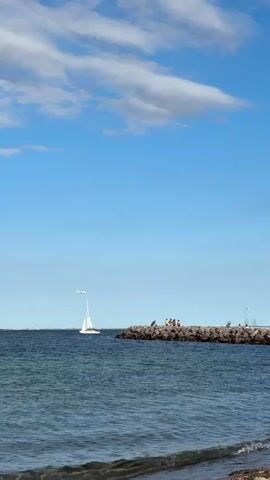 The video shows a wide shot of a body of water under a blue sky with scattered clouds. In the distance, a white sailboat with its sail up is visible on the left side of the frame. To the right of the sailboat, there is a stone jetty. Several people are standing and sitting on the jetty. A small white airplane is flying in the sky above the jetty. The water in the foreground has small waves breaking on a sandy shore. The overall scene appears to be a calm day at the beach or coast.