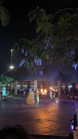 At night, two women in light-green skirts and headbands perform with fire batons on a stone-paved stage in front of an audience. They twirl and toss the lit batons as part of the performance. Behind them is a building with people watching from the porch. A tiki-like rock formation stands to the left of the stage. Lush green tree branches frame the top of the shot, with outdoor lighting illuminating the scene.