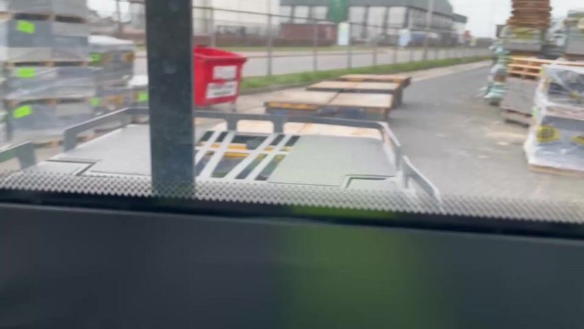 The video shows a person driving a forklift in an industrial area. The view is from inside the forklift cabin, looking through the windshield. The initial shot is focused on the lower part of the windshield, where a perforated black border is visible. Beyond the windshield, there are stacks of materials, possibly construction supplies, wrapped in plastic and sitting on pallets. There's also a red container further away and some flatbed trailers in the background.

The camera then shifts upward and to the right, showing a view of a road, grassy verge and perimeter fence through the side window of the forklift. 

Finally, the camera angle shifts again, capturing a hand with a light purple fingernail driving the forklift. The hand is holding the steering wheel. A switch with a green light is visible on the dashboard. The wipers are off, and the road ahead is paved. The forklift appears to be in motion.