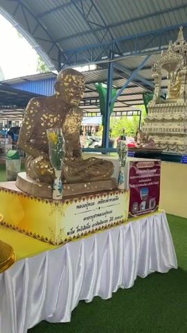 A video shows an outdoor Buddhist shrine area. The first shot focuses on a statue of a monk covered in gold leaf, seated on a platform draped in white and gold fabric. Two vases with flowers are placed in front of the statue. The second shot pans to the right, revealing a large, ornate silver structure with a Buddha statue inside. Green artificial turf covers the ground. In the background, there are trees covered in netting and various buildings. The third shot shows a side view of the area, with more of the buildings and a glimpse of a white car. The final shot shows the front of a building with a dark blue roof and wooden pillars. A large golden elephant statue stands on the left, and the entrance to the building reveals several Buddha statues inside. People are visible entering and moving within the building.