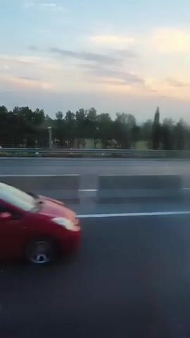 The video shows a view from a vehicle traveling on a highway. A red car is seen in the foreground, traveling in the same direction. In the background, there is a line of trees and a cloudy sky, suggesting it is either sunrise or sunset. Another silver car is visible on the highway in the middle distance. The highway has multiple lanes separated by concrete barriers. The lighting suggests it is late afternoon or early evening.