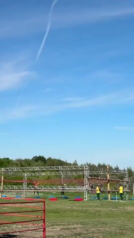 The video shows a ground level shot of a recreational outdoor obstacle course with a team of young people participating in the event. The event takes place on a clear, sunny day under a bright blue sky. The course consists of silver scaffolding, red netting, red and blue floor mats and a final obstacle of tree logs with wooden handholds, assisted by event staff. The young people are dressed in athletic gear and seem to be moving from one event to the next. Beyond the obstacle course a green tree line is visible. In the sky, a single contrail from an aircraft is visible.