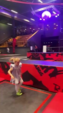 A little girl with blonde hair in pigtails runs and jumps at a trampoline park. She is wearing a blue dress, white tights, and bright green socks. The trampoline is black and red and the walls feature graphic designs in red and black. The girl jumps toward the wall where there are lighted targets and hits some of them. Several other people can be seen in the background, and the lighting in the building appears to be mostly blue and purple.