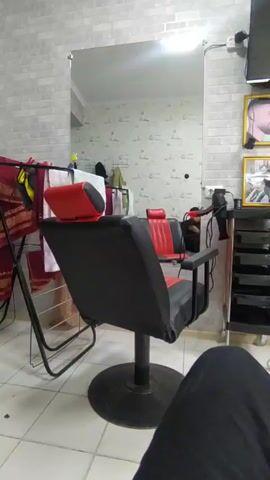 A stationary, medium shot shows a barber shop setting. A black barber chair with red accents sits in the center of the frame, facing a large mirror. Reflected in the mirror is a view of the shop's interior, including a patterned wall, towels hanging on a rack, and other barbering tools. To the right of the chair, a black rolling cart with drawers holds a hairdryer and other items. The floor is tiled in a light color. A portion of a person's leg, clad in black pants, is visible in the lower right corner, suggesting someone is present in the room. The lighting appears to be bright and even, possibly from overhead lights.