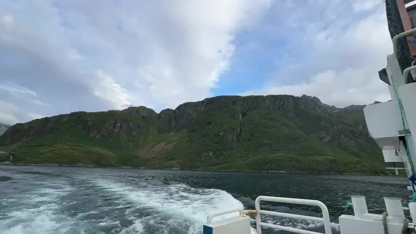 The video shows a view from a moving boat on a body of water. The boat is leaving a wake behind it. In the distance, there are green, tree-covered mountains with rocky peaks. A large bridge spans across the water, connecting two landmasses. The sky is cloudy with patches of blue visible. The overall scene is of a scenic, natural landscape.