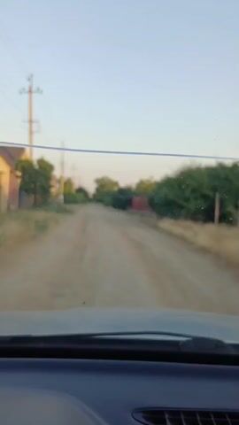 The video shows a view from inside a car looking out through the windshield. The car is traveling on a dirt road in what appears to be a rural area or a small town. The road is unpaved and has some ruts and unevenness. On either side of the road are houses and trees. Power lines and utility poles are visible along the road. The sky is clear and the lighting suggests it is either early morning or late afternoon. The dashboard of the car is visible in the foreground.