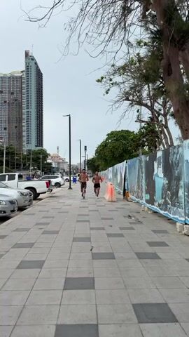 Two shirtless men are running on a paved sidewalk. The man on the left is wearing camouflage shorts and white socks with black and pink athletic shoes. He has a beard and is flexing his bicep. The man on the right is wearing black shorts and black athletic shoes. He has short dark hair and is looking to his right. They are running towards the camera. In the background, there are cars parked along the side of the road, and a tall building in the distance. To the right of the sidewalk, there is a fence with large images on it, and trees. The sky is overcast.