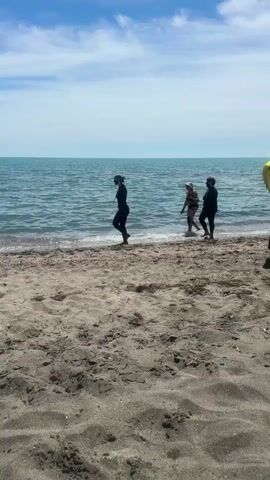 Three people walk along the shoreline. Two of the individuals are wearing dark full-body suits, while the third person is wearing a bikini and a hat. The water is calm and blue, with gentle waves lapping at the shore. The beach is sandy and appears to be made up of small pebbles and shells. The sky is clear and blue with some scattered clouds. The video is shot from a slightly elevated position, looking down at the scene.