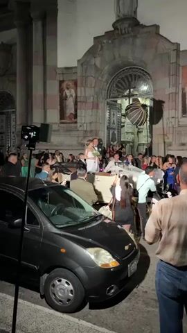 A wedding party exits a church. The bride and groom are in the foreground, posing for photos in front of the church doors. A wedding reception is underway, with guests dressed in formal attire. The bride is wearing a white wedding dress, and the groom is wearing a suit. A black car is parked in front of the church, and people are taking photos of the wedding party. The church is made of stone, and there are paintings on the walls. The wedding party is in high spirits.