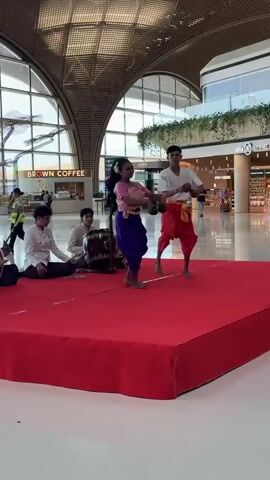 This is a video of a traditional Cambodian dance performance. Two young dancers, a girl and a boy, are on a red stage, performing in front of an audience in what appears to be an airport. The girl is wearing a pink top and a purple sarong, while the boy is wearing a white shirt and orange pants. Behind them, three musicians are playing traditional Cambodian instruments. The environment is a large, modern indoor space with a high ceiling, large windows, and shops in the background. The dancers are moving gracefully, with the girl leading the dance. The musicians are playing a drum, a string instrument, and another instrument that is not clearly visible. The overall atmosphere is festive and cultural.