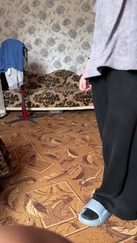 The video shows a person standing in a room with floral wallpaper. The person is wearing a grey t-shirt, black pants, and blue slippers. The person is holding a phone and recording. The room has a sofa and a fan. The floor is carpeted. The person walks away from the camera.