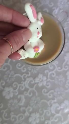 This is a close-up, overhead shot of a mug of coffee or hot chocolate with a marshmallow bunny floating in it. The bunny is white with pink accents on its ears and paws, and has a small pink flower and green leaf decoration on its chest. The mug appears to be made of glass. The background is a light-colored tablecloth with a floral pattern. The image conveys a sense of whimsy and springtime, possibly related to Easter. There is no visible text in the image. The lighting suggests it could be daytime, indoors.