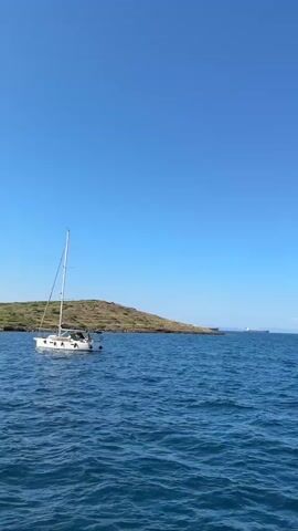 A wide shot of a sailboat on blue water shows a small, hilly island on the left. The boat is traveling left to right, and in the background, a large ship is visible, with other distant land features. The sky is clear and blue.
