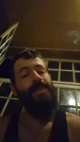 A man with a beard and dark hair, wearing a black tank top, is seen from the chest up in a medium close-up shot. He is looking directly at the camera and exhaling smoke, possibly from a cigarette. The background shows a window with multiple panes and an open section above. The lighting suggests it is nighttime or indoors with artificial light. The man's expression is neutral.
