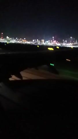 The video depicts a nighttime scene from the window of an airplane. The view shows the wing of the plane and the airport runway illuminated with lights. In the distance, a city skyline with scattered lights is visible against the dark sky. The video appears to be taken during the plane's takeoff or landing process.