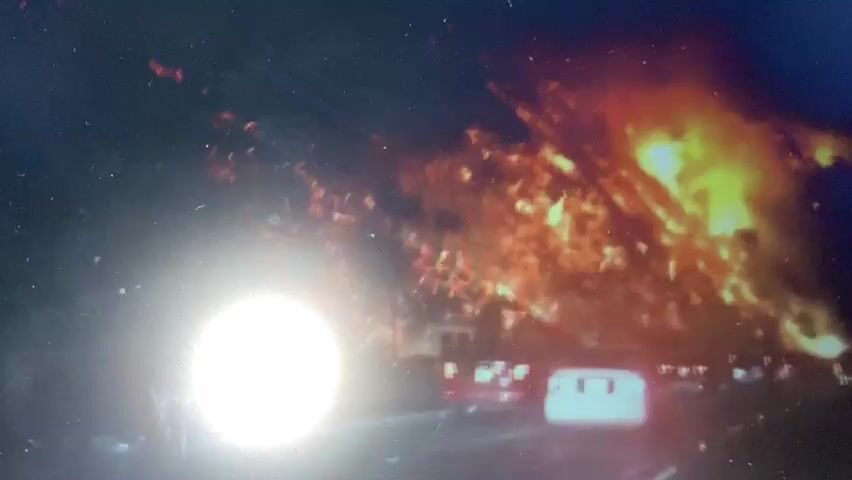 The video shows a bright white light emanating from the left side of the screen. To the right of the light, there is a large, chaotic blaze with flames and smoke. In the background, there appear to be buildings and vehicles, including a white car. The overall scene suggests an intense event, possibly an explosion or a major fire, occurring at night. The lighting conditions are extreme, with the bright light overwhelming some details of the scene.