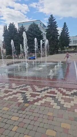 The video shows an outdoor fountain on a sunny day. The fountain is a rectangular shape with multiple jets of water spraying upwards. The area around the fountain is paved with colorful interlocking bricks in a geometric pattern. In the background, there are several tall evergreen trees and a multi-story building. A young girl in a pink shirt and shorts can be seen to the right, walking on the wet pavement.