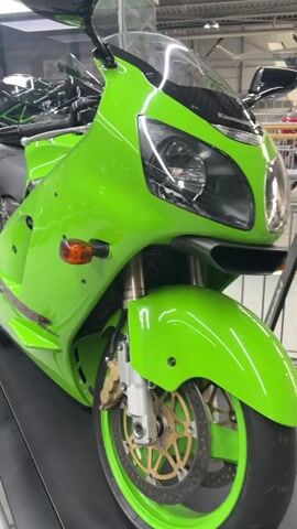 The video shows a close-up view of a green Kawasaki Ninja motorcycle displayed indoors. The camera pans across the bike, highlighting its sleek design and vibrant color. The motorcycle is equipped with a black seat, chrome exhaust pipes, and gold accents on the wheels. In the background, other motorcycles and some aircraft can be seen, suggesting the setting might be a museum or a showroom. The lighting is bright, and the overall atmosphere is clean and well-maintained. No people are visible in the frame.