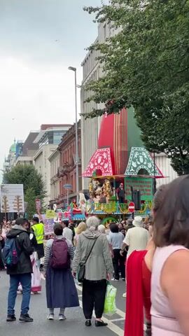 The video shows a street scene with a parade. In the foreground, a woman with long brown hair tied back is looking away from the camera, wearing a red garment. Behind her, a crowd of people are walking down the street. A large, colorful float, resembling a temple or chariot, is moving in the distance, adorned with red, green, and gold decorations. Figures are visible on the float, and some people in the crowd appear to be holding signs. The environment appears to be an urban setting with buildings on either side of the street and trees lining the roadway. The sky is overcast.