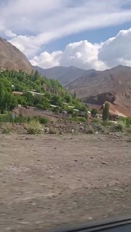 The video shows a mountainous landscape with a small village nestled on a hillside. The mountains are mostly barren, with some patches of greenery, and the sky is blue with fluffy white clouds. The village consists of a few buildings scattered among trees. The foreground is a dirt road or clearing. The video appears to be taken from a moving vehicle.