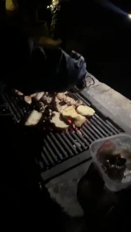 A person is cooking food on a grill at night. The grill is outdoors and appears to be made of metal. There are several slices of bread and some pieces of meat on the grill. The coals underneath the grill are glowing red. In the background, there are some lights strung up and what appears to be some bags of groceries or supplies. The overall atmosphere seems to be a casual outdoor gathering.