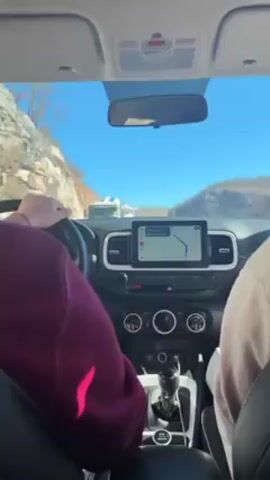 The video shows the interior of a car, filmed from the back seat looking forward. Two people are visible in the front seats; the person in the driver's seat is wearing a maroon sweater and is seen gripping the steering wheel, while the passenger is wearing a tan sweater and can be seen using their phone. The car is traveling on a highway in a mountainous area on a clear day with a blue sky. A GPS navigation system is mounted on the dashboard, displaying a route map. Ahead of the car, a large white truck is traveling in the same direction. The scenery includes rocky hillsides and snow-capped mountains in the distance.