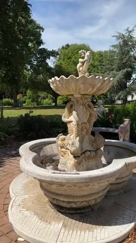 A stone fountain sits on a brick patio surrounded by a green lawn and trees. The fountain is ornate, with carvings of dragons and cherubs. It is a sunny day, and the sky is blue with some clouds. There are people walking in the background on the lawn. The fountain is not currently running water.
