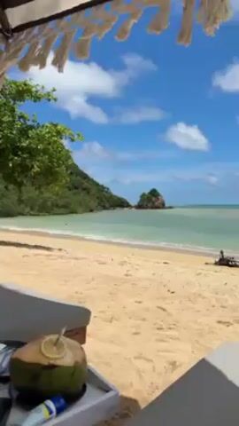 The video shows a person lying on a beach lounger with a coconut drink on a tray next to them. The beach is sandy with clear turquoise water and green hills on either side. There are a few people on the beach, including two children who appear to be playing in the water. The sky is blue with puffy white clouds. The camera angle is from the person's point of view.