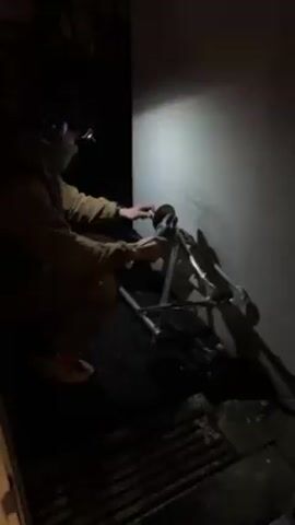 In a dimly lit room, a person wearing a headlamp, goggles, and a mask is crouched and using a cutting tool on a piece of metal. The person is also wearing a brown shirt, a watch, and distinctive patterned shoes. The cutting tool is mounted on a metal frame that appears to be a modified walker or similar mobility aid. Sparks are visible as the tool cuts through the metal, and there are metal shards and debris scattered on the floor. The scene is confined, with a light-colored wall behind the person and dark areas filling the periphery.