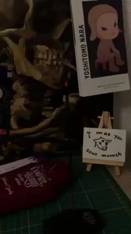 The video presents a still life arrangement on a cutting mat. At the back, a painting depicting a skull dominates. To its right, a postcard featuring art by Yoshitomo Nara leans against the wall. In front of the painting, an incense stick burns from a holder that sits on a small tombstone engraved with "RIP". Adjacent to the incense, a coffin-shaped box labeled "Vampire Blood" rests. A tiny easel displays a drawing of a cow, accompanied by the phrase "I moo you sooo muuch". The scene appears to be staged with a gothic or whimsical theme.