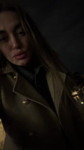 At night, a woman in a brown coat and dark turtleneck films herself as she walks outside. The video then abruptly shifts its focus to a statue of a man on a horse situated on a stone platform. The background reveals a city scene with streetlights and building lights. The video quickly becomes shaky and out of focus, blurring the cityscape.