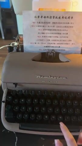 A close-up shot shows a hand with manicured nails typing on a vintage Remington Quiet-Riter typewriter. The typewriter is a light brown or beige color, with black keys and a silver carriage. A piece of paper with Chinese text is loaded into the carriage, and the paper appears to be a set of instructions. The hand is positioned over the keyboard, with fingers poised to strike the keys. The background is blurred, but it appears to be an indoor setting with warm lighting. The video captures the action of typing, with the keys being pressed and the paper moving forward with each stroke. The sound of the typewriter is not included in the video.