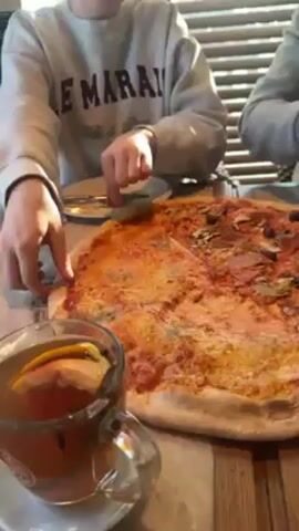 A person wearing a light gray sweatshirt with the word "LE MARAIS" printed on it pulls a slice of pizza from a large pizza on a table. The pizza is topped with cheese, tomato sauce, and other ingredients. A cup of tea with a slice of orange is also on the table. The person then puts the slice of pizza in their mouth. Another person wearing a gray shirt is visible in the background. The table is made of wood.