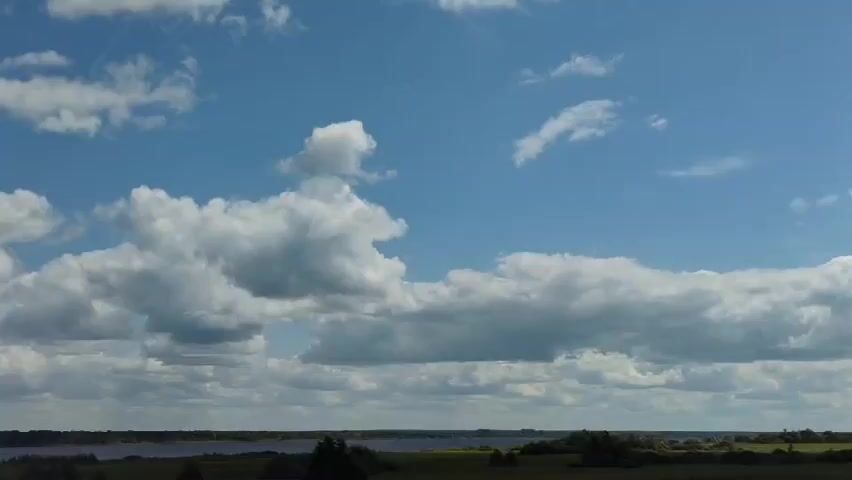 A video shows a wide shot of a landscape on a bright, partly cloudy day. A body of water, possibly a lake or river, stretches horizontally across the frame in the middle ground, bordered by green fields and trees. The sky above is a clear blue, dotted with fluffy white cumulus clouds of varying sizes and densities. The camera appears to be stationary, offering an unobstructed view of the serene natural scene. There are no people or discernible objects in the foreground or middle ground. The overall impression is one of a peaceful, natural environment.