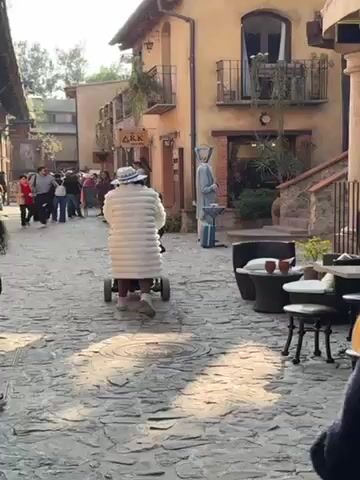 The video shows a person wearing a white, textured coat and a white hat with a patterned brim walking away from the camera and pushing a stroller along a cobblestone street. The person is also wearing brown pants and white boots. The street is lined with buildings that look like they are in a Mediterranean village, with stone and stucco facades and wrought iron balconies. There are other people walking around in the background. There is a sign for "ARK" above a shop. A mannequin stands outside a store to the right.