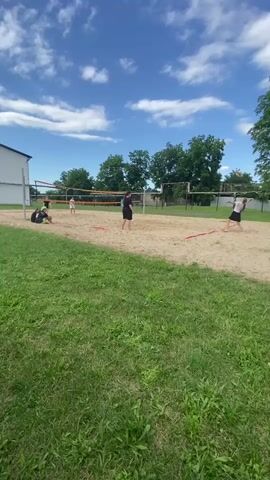 A group of people are playing volleyball on a sand court. There are four people visible on the court, two on each side of the net. One person is in the process of serving the ball, while the others are positioned to receive or play the ball. The court is marked with red lines. The game is taking place outdoors, with a grassy area in the foreground and trees and a building in the background. The sky is blue with white clouds, suggesting pleasant weather. The sun appears to be high in the sky, indicating it is daytime, likely afternoon. The overall mood appears to be casual and recreational. No text is visible in the media. The location context suggests this is Mostovskoy, Russia.