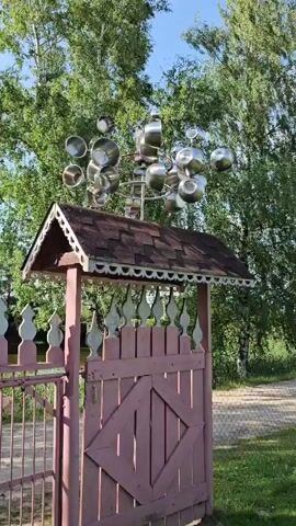 The video shows a kinetic sculpture made of pots and pans that is spinning in the wind. The sculpture is mounted on top of a decorative gate structure. The gate is made of wood and painted pink and white. It has a pitched roof with shingles. The background is filled with green trees and a bright sky, suggesting an outdoor setting during daytime. The sculpture itself consists of multiple metallic pots and pans of various sizes arranged in a circular pattern on a central pole. The movement of the pots and pans creates a dynamic visual effect. The overall scene appears to be peaceful and rustic.